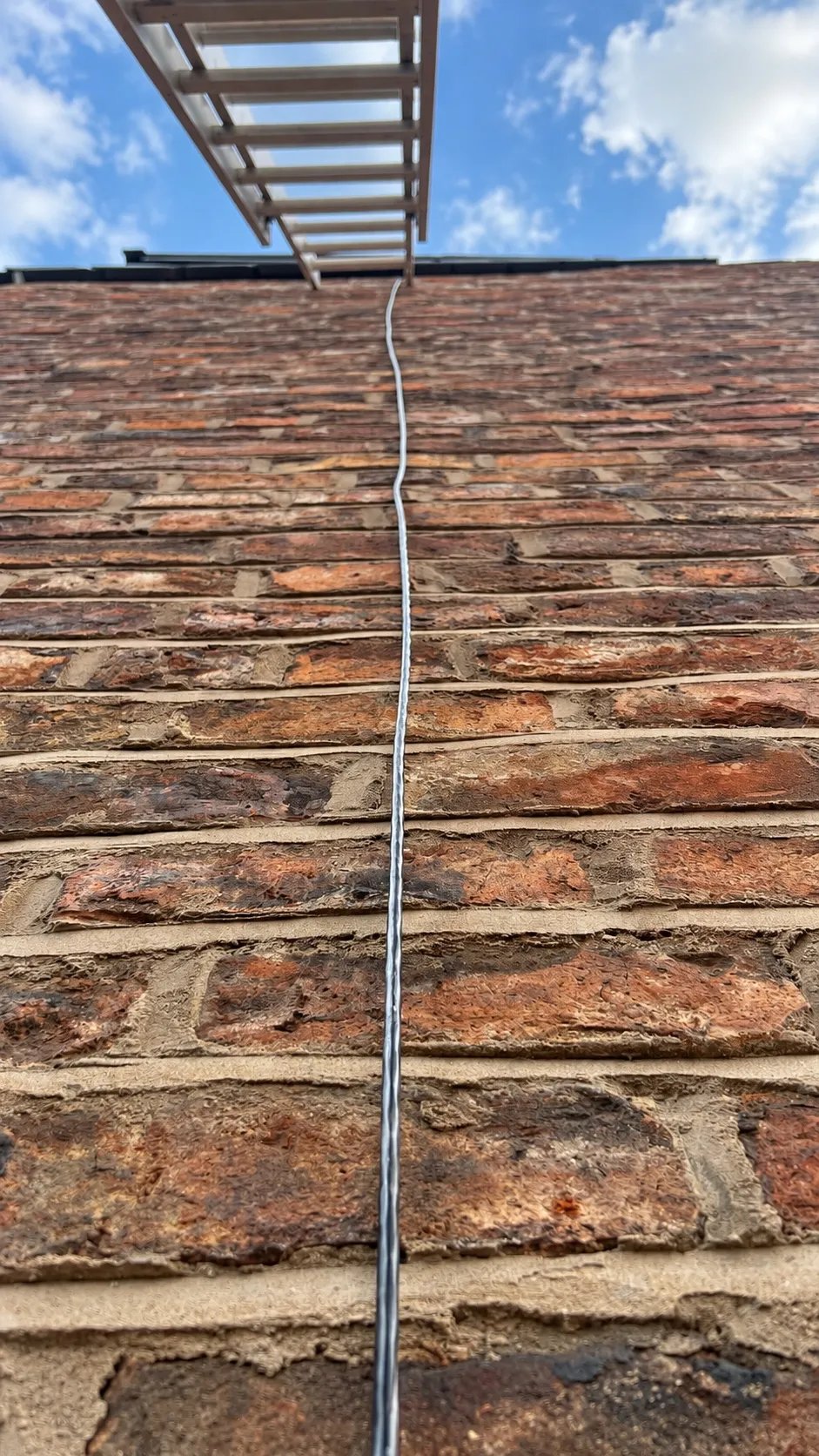 Looking straight up a brick gable wall: one thin black network cable runs up the brickwork to a ladder at the roofline